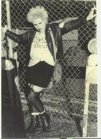 A black-and-white photo of a person with spiky hair, wearing a leather jacket, a t-shirt with the words 'no idea' printed on…