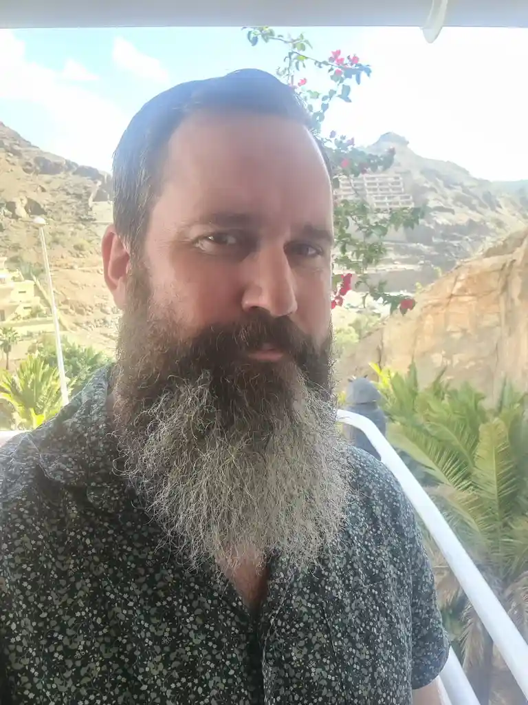 A man with a long, graying beard and dark hair looks directly at the camera from a balcony. Behind him, a sunny, rocky…