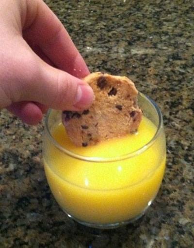 A hand holds a chocolate chip cookie partially submerged in a glass of orange juice on a speckled countertop. The overlaid…