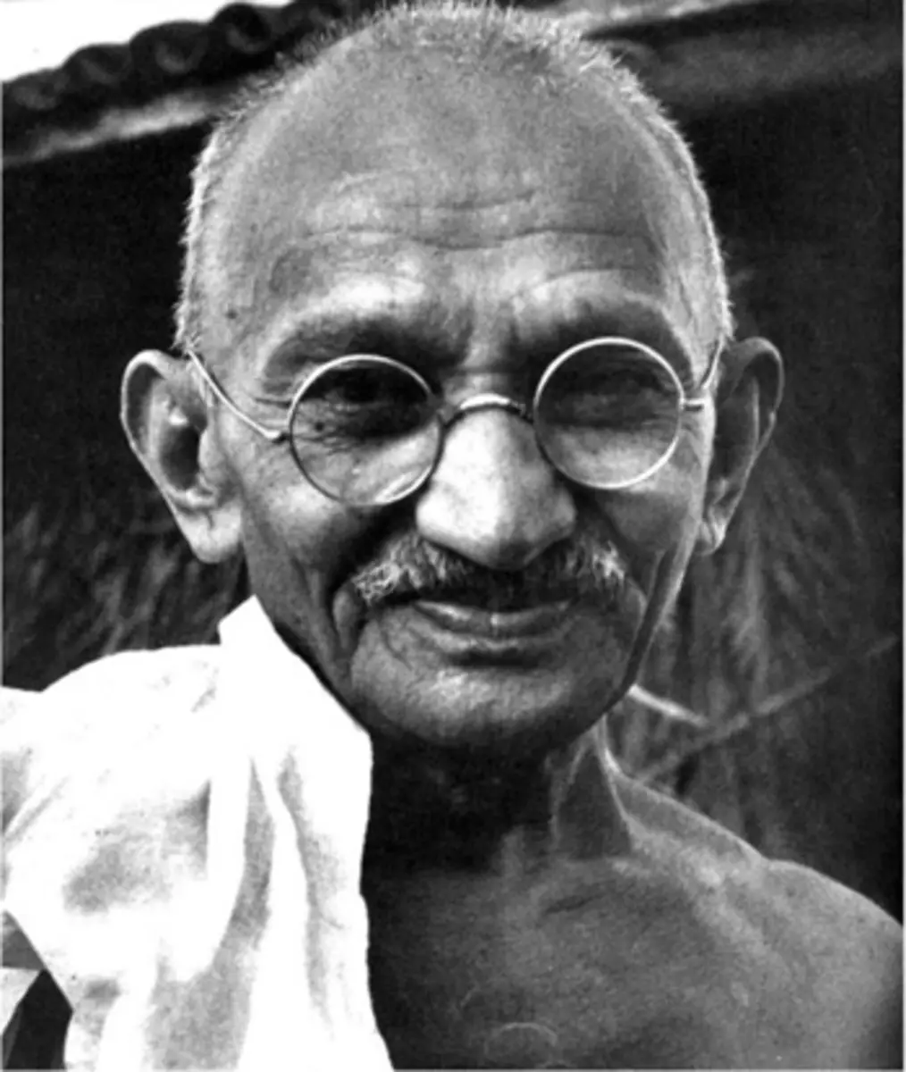 A black-and-white photograph of Mahatma Gandhi smiling, wearing round glasses and a white cloth draped over his shoulder…