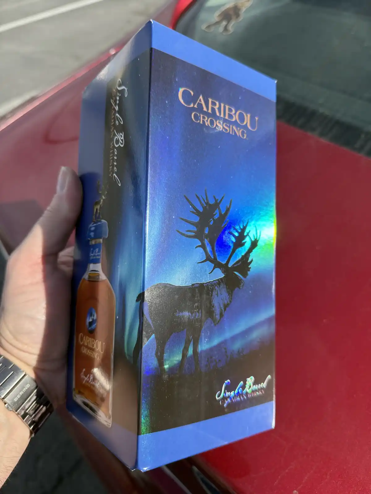 A hand holds a blue box of Caribou Crossing Single Barrel Canadian Whisky against a red car. The box features a silhouette…
