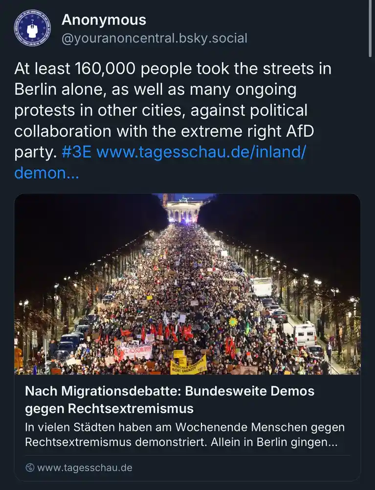 A high-angle nighttime photo shows a massive crowd of protesters filling a wide Berlin street, heading toward a brightly lit…