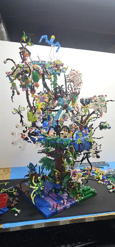 A complex, chaotic LEGO tree sculpture covered in colorful minifigures, spiders, and foliage, with scattered LEGO pieces on…