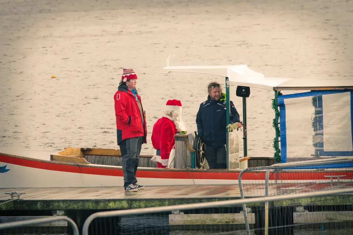 A man dressed as Santa Claus stands on a small boat docked at a pier, facing another man in a red jacket and striped hat. A…