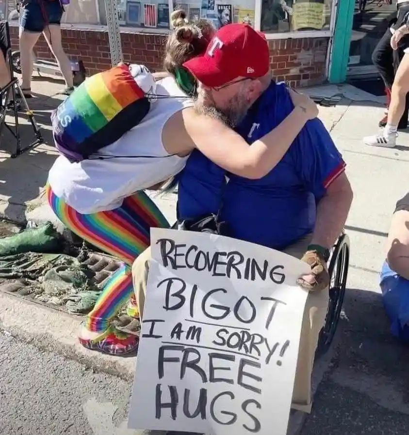 A man in a wheelchair wearing a red baseball cap and blue shirt is being hugged by a person in rainbow-colored clothing and…