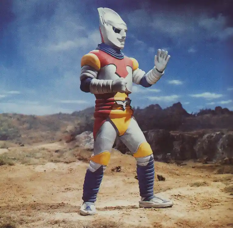 A full-body shot of the Ultraman character Ultraseven stands in a desert-like landscape under a blue sky with clouds. He…
