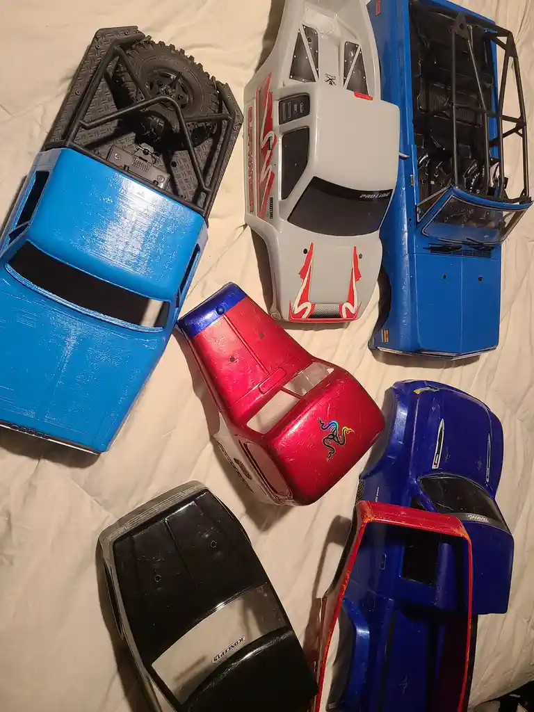 Several disassembled radio-controlled truck bodies in various colors—blue, red, black, and white—are laid out on a white…