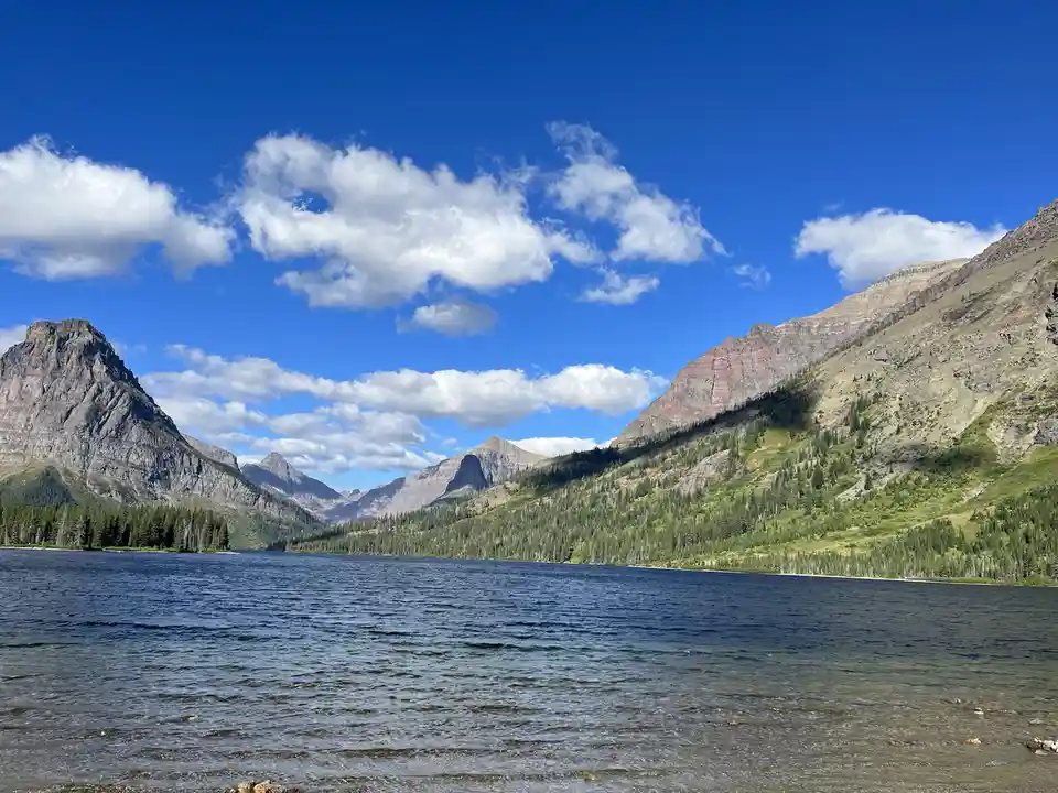 A scenic photograph of a calm lake surrounded by towering, rugged mountains under a bright blue sky with scattered white…