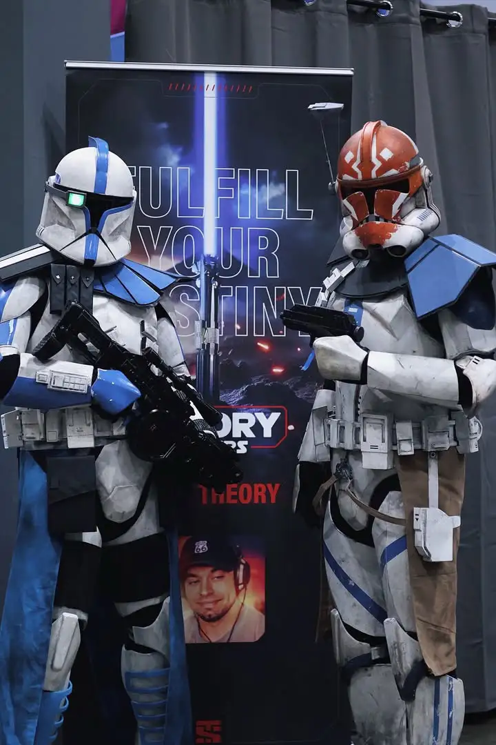 Two people in Star Wars clone trooper costumes stand in front of a promotional banner. The banner features the text 'FULFILL…