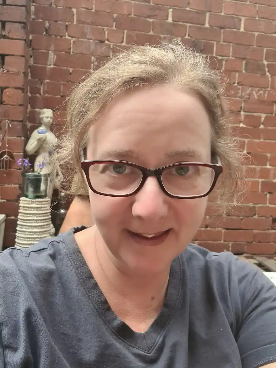 A woman with glasses and messy hair smiles at the camera in front of a brick wall. Behind her is a small statue and a beaded…
