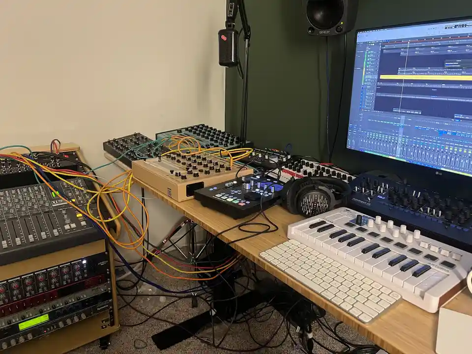 A cluttered music production studio desk is filled with numerous electronic music equipment including synthesizers, a mixer…