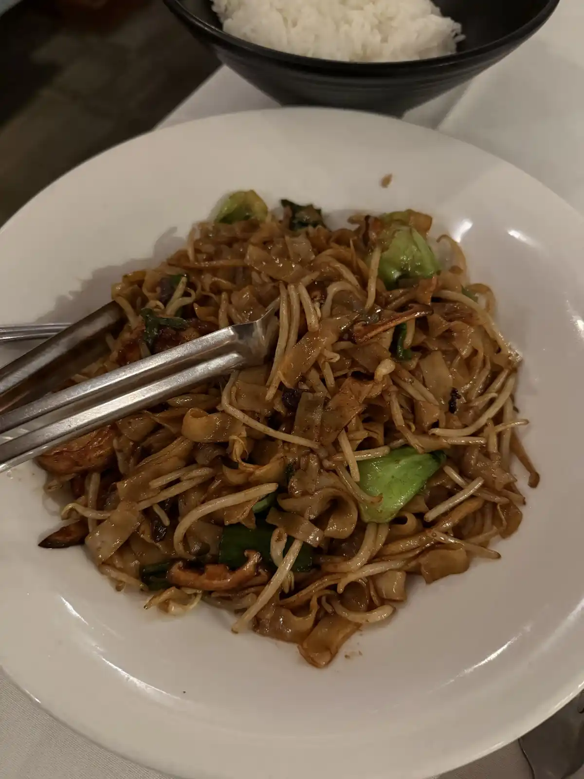 A close-up photo of a plate of stir-fried rice noodles with vegetables and mushrooms, with metal tongs resting on top. In…