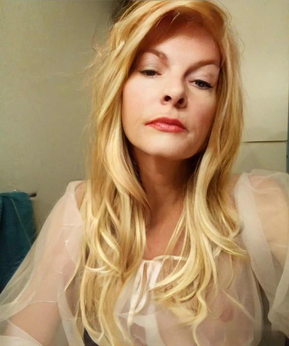 A woman with long blonde hair and red lipstick looks directly at the camera with a tired, unimpressed expression while…