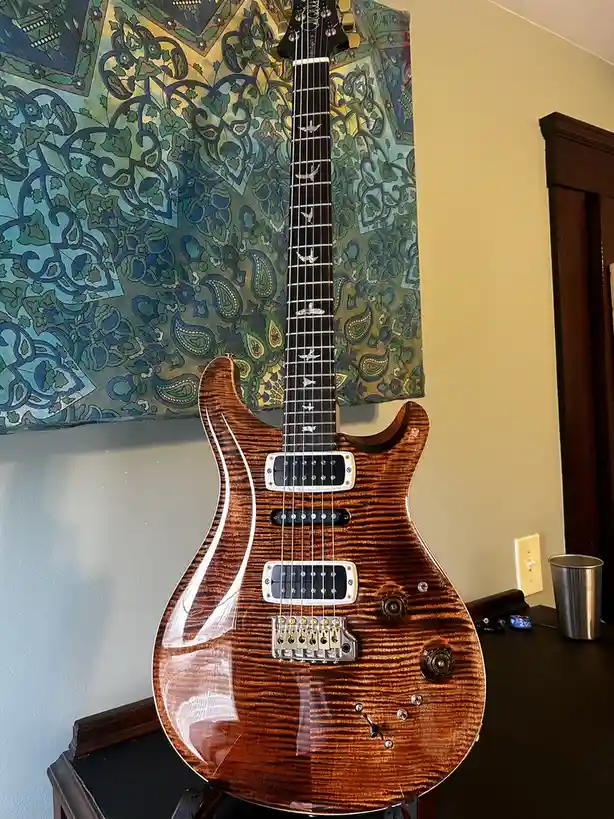 A glossy, wood-grain electric guitar with white pickups and gold hardware stands upright on a dark wooden stand against a…