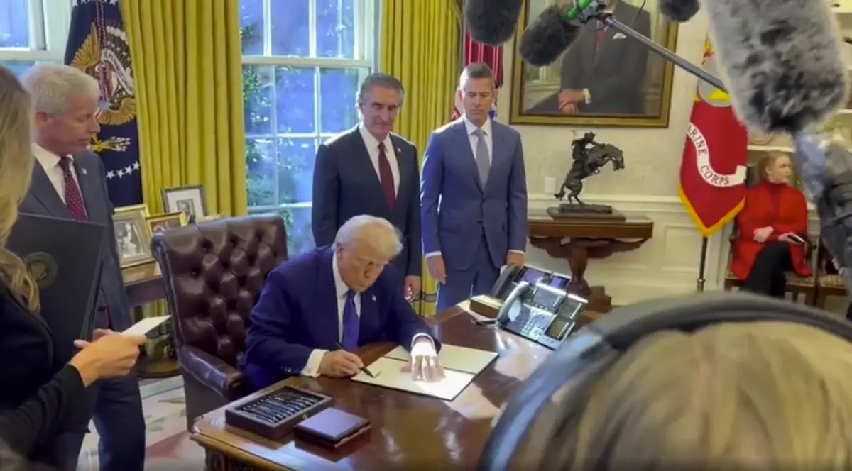 A man in a blue suit sits at a large wooden desk in an ornate office, signing a document while several people in suits stand…