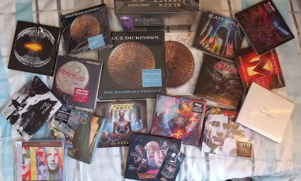 An overhead shot displays a large collection of new, sealed heavy metal music albums spread across a patterned bedspread…