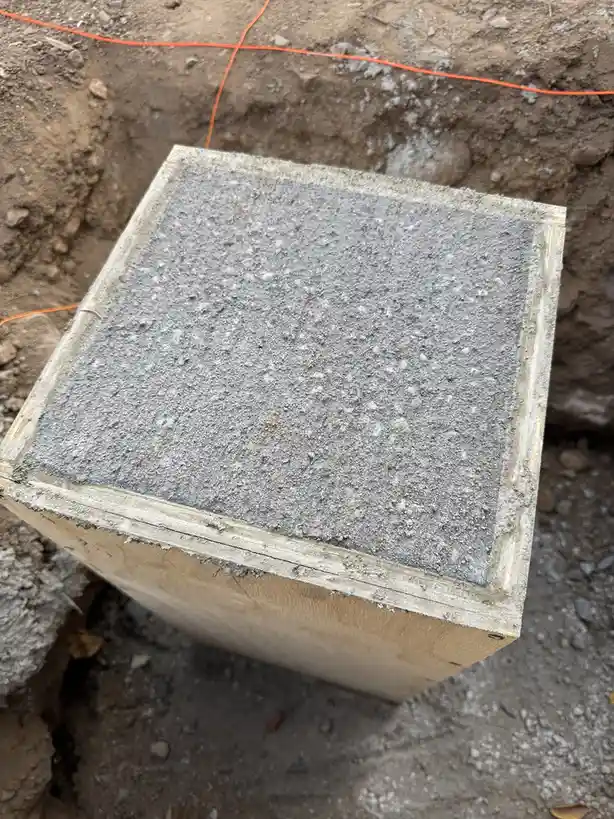 A close-up photo shows a freshly poured, gray concrete block still inside a wooden form, set in dirt with an orange…