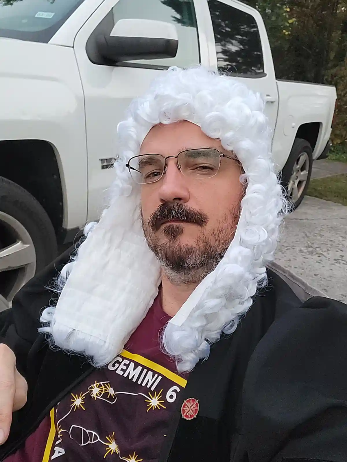A man with glasses and a mustache wears a white, curly judge's wig and a black robe over a maroon t-shirt that says 'GEMINI…