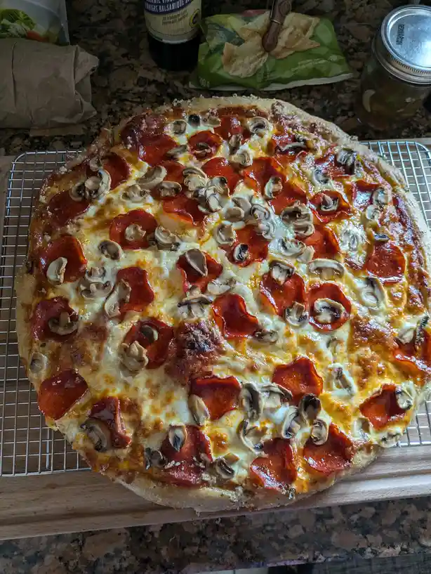A freshly baked pepperoni and mushroom pizza sits on a wire cooling rack atop a wooden board, surrounded by kitchen items…