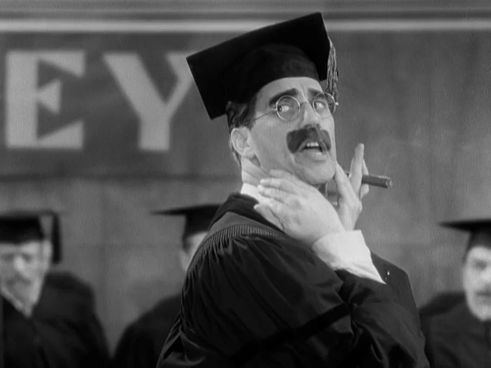 A black-and-white image shows a man in a graduation cap and gown, wearing round glasses and a mustache, holding a cigar…