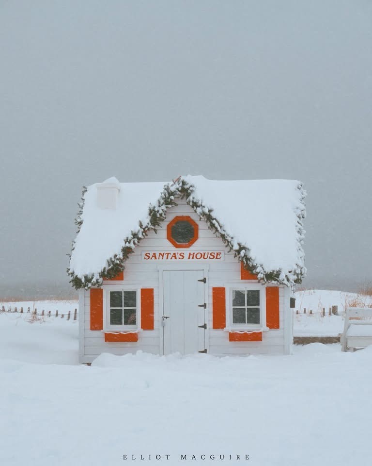 A small white wooden house with orange trim and a snow-covered roof, decorated with evergreen garland and a circular window…
