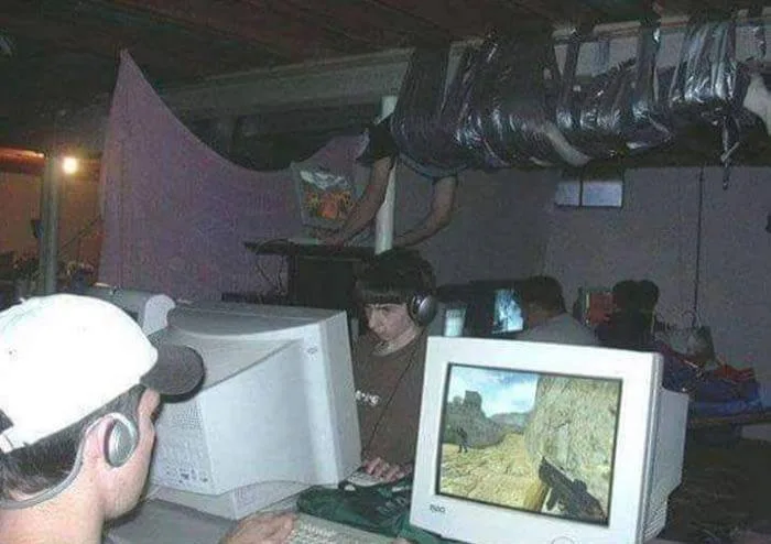 A dimly lit, cluttered room with multiple people intensely focused on old CRT computer monitors displaying a first-person…