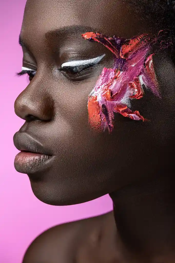A close-up profile of a Black woman with flawless skin against a pink background, featuring a sharp white winged eyeliner…