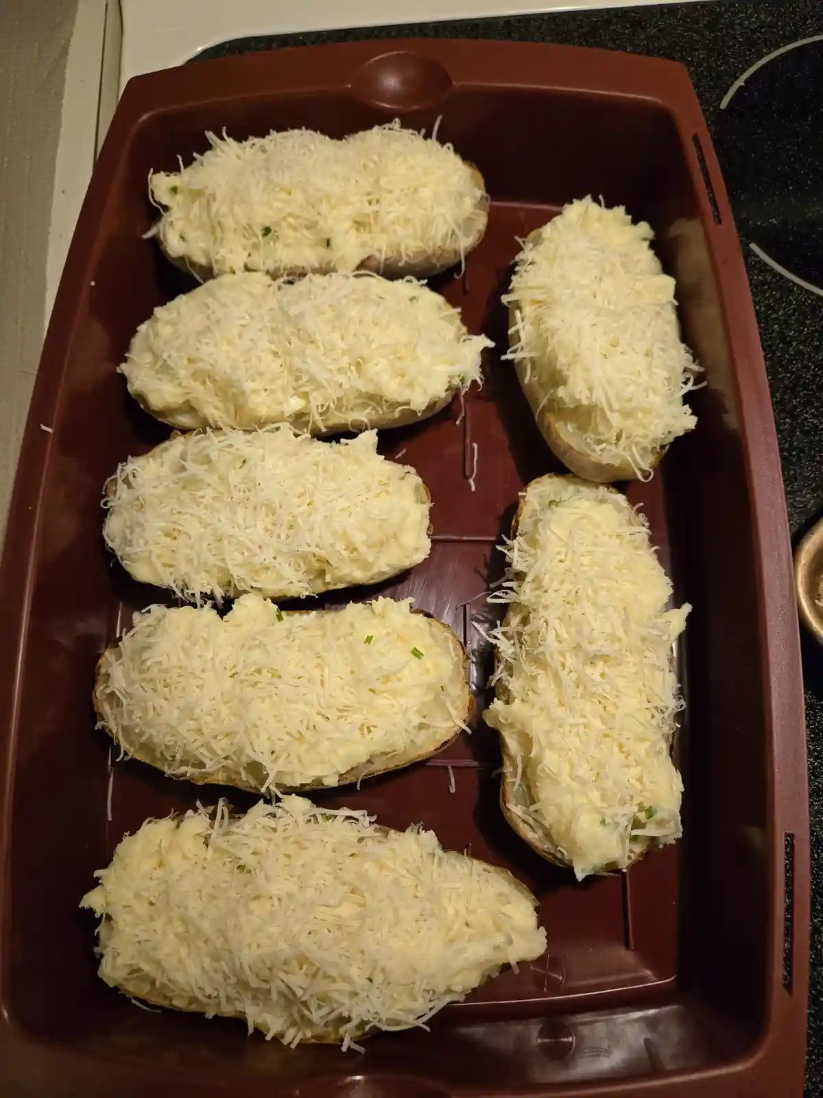A brown baking tray holds six baked potato halves, each generously topped with creamy mashed potato filling and a thick…