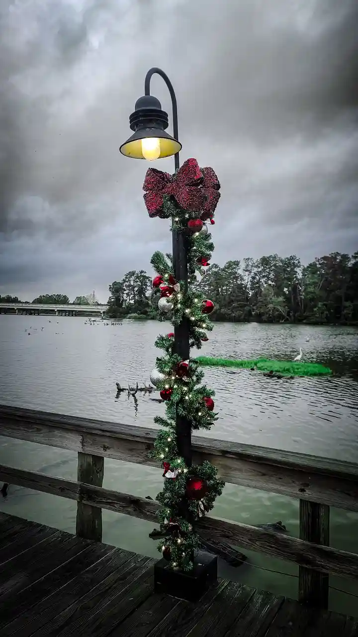 A black lamppost on a wooden pier is heavily decorated with Christmas garlands, red and silver ornaments, and a large…