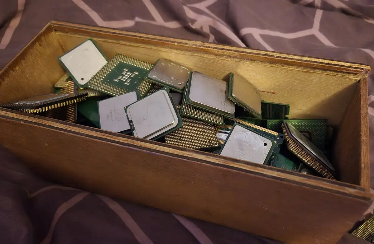 A wooden box filled with various old computer processors and RAM sticks sits on a patterned fabric surface. The overlaid…