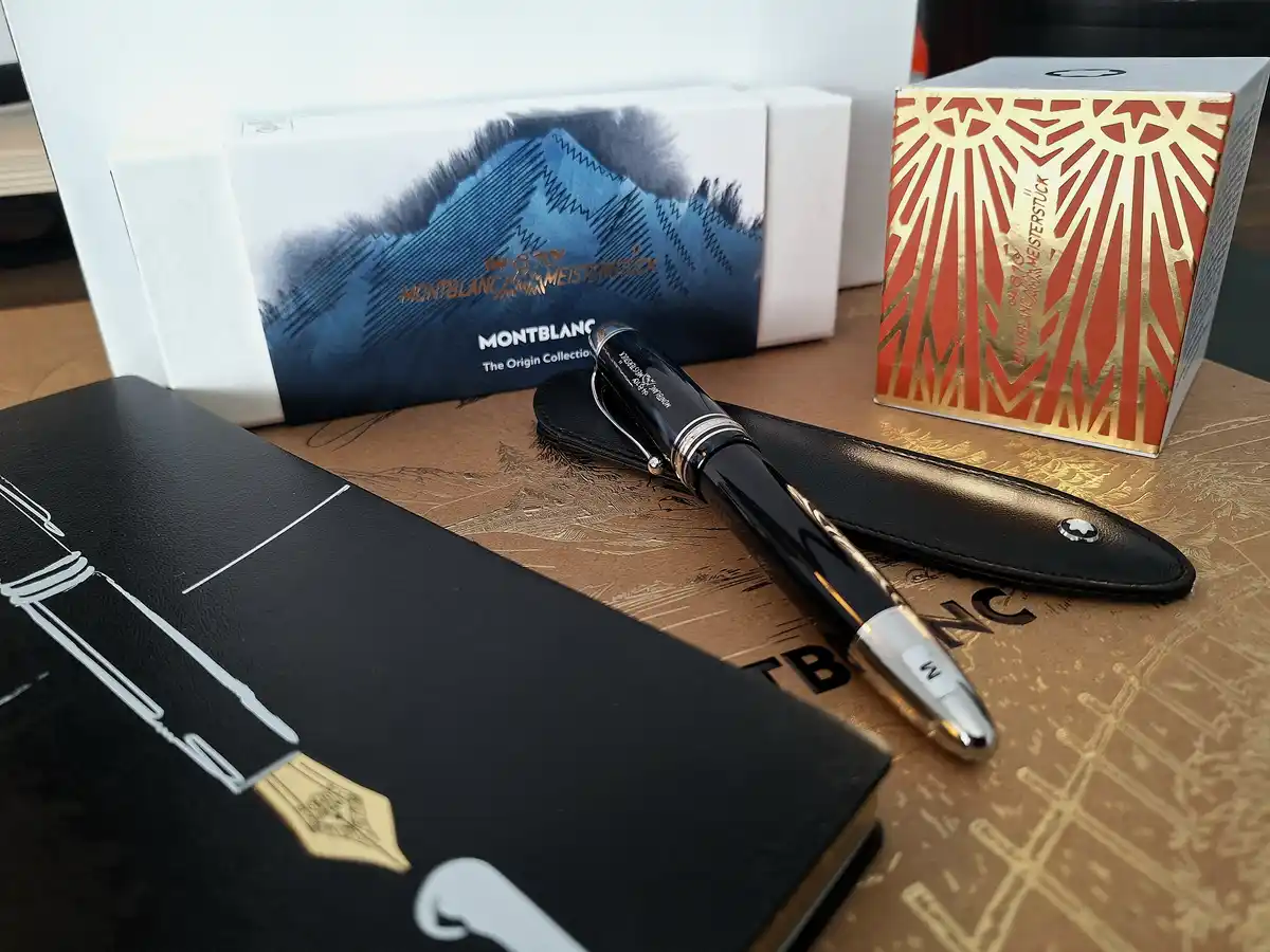 A high-end Montblanc Meisterstück fountain pen rests on a brown patterned surface next to its black leather case and two…