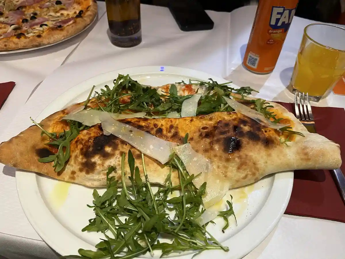 A close-up photo of a folded, half-moon shaped pizza on a white plate, topped with arugula and shaved cheese, with another…