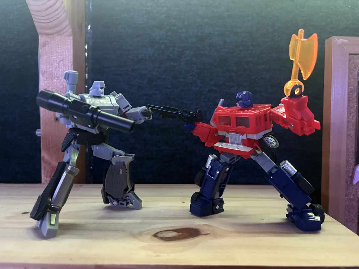 Two transformer action figures, one grey and one red, are facing each other with weapons drawn. The grey figure holds a…