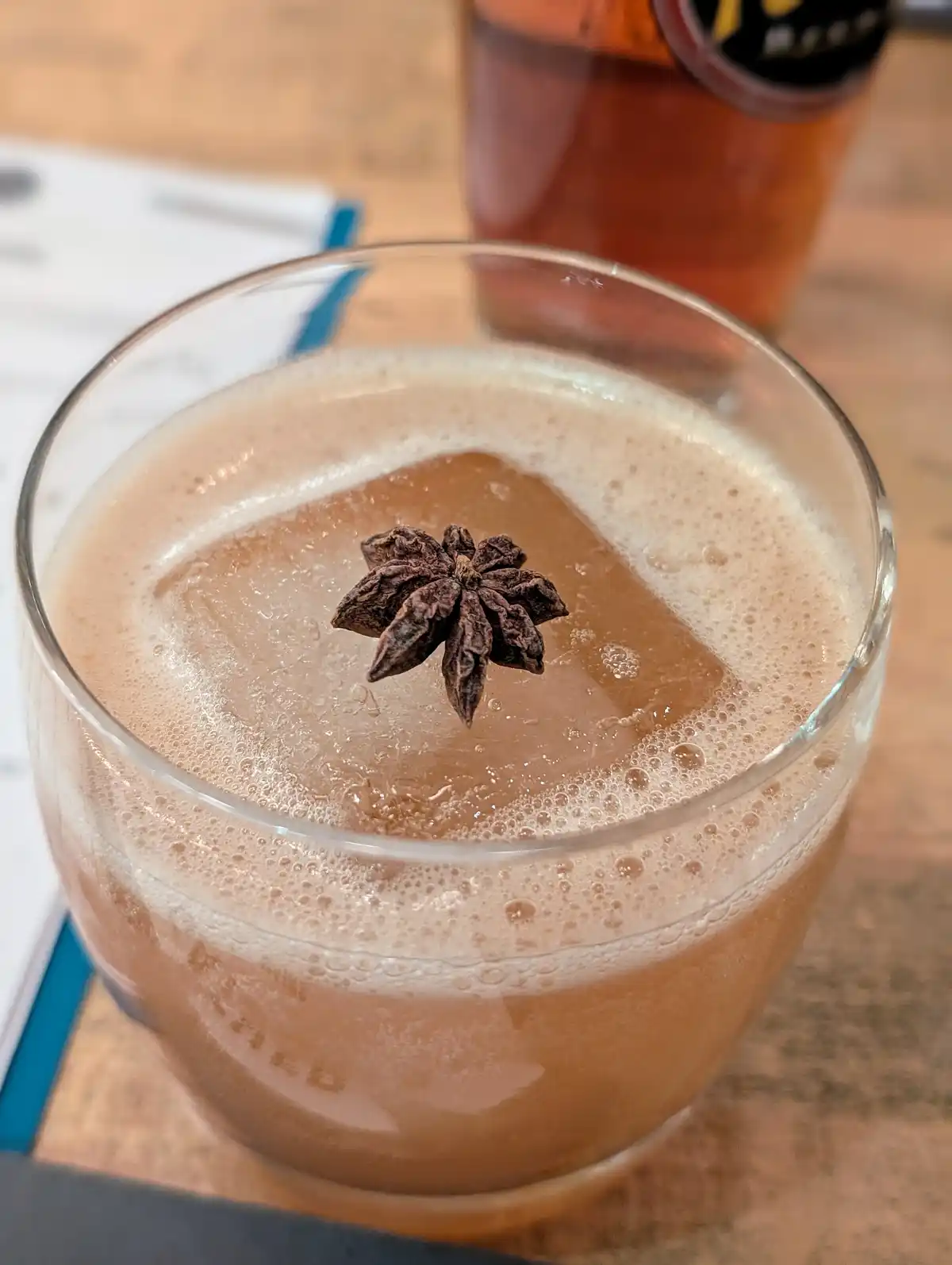 A close-up photo shows a glass of amber-colored cocktail with a large ice cube and a whole star anise floating on top, set…