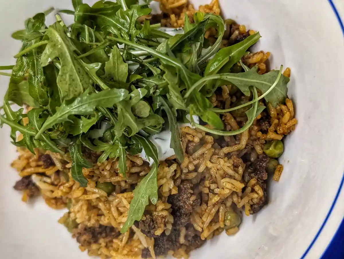 A close-up photo of a white bowl containing a dish of brown rice mixed with meat and peas, topped with a generous pile of…