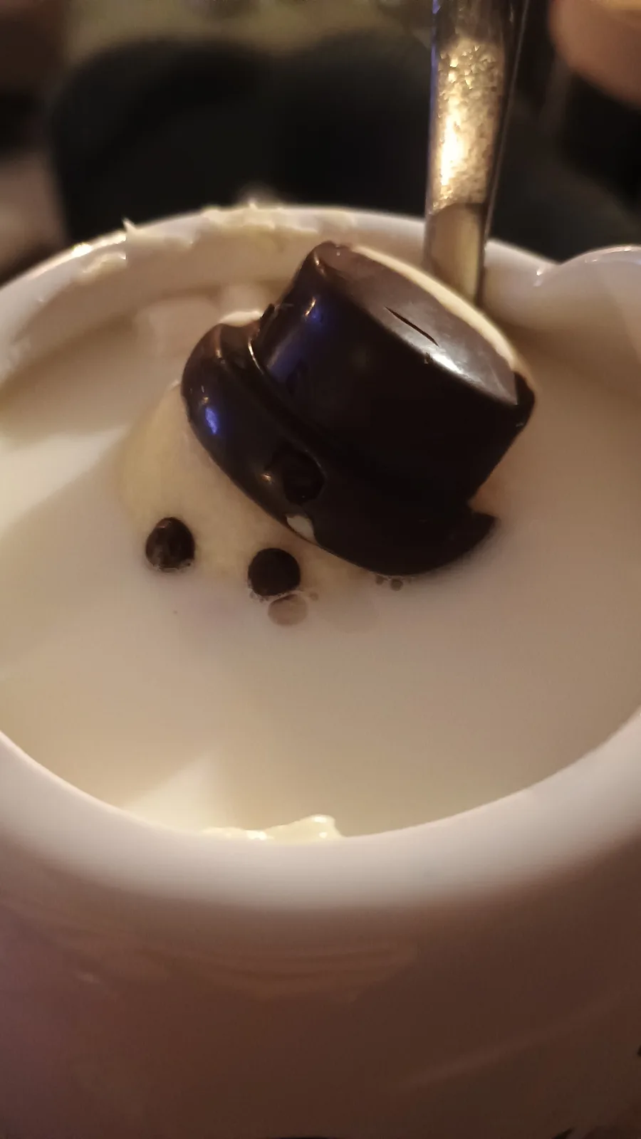 A close-up photo of a dessert in a white bowl, featuring a glossy chocolate dome with two small chocolate dots below it…