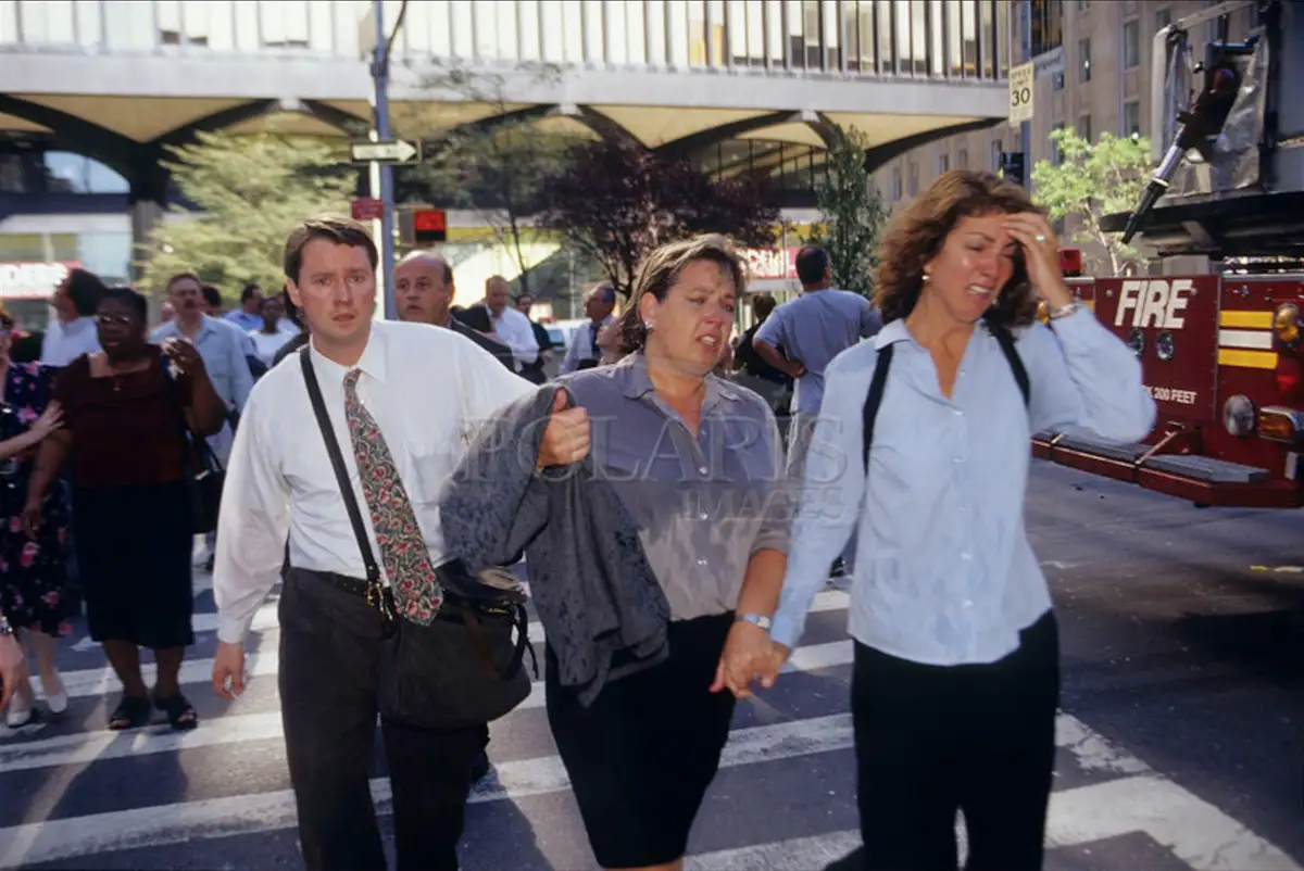 A photo shows three people walking across a city street crosswalk, visibly distressed. A woman in the center appears to be…