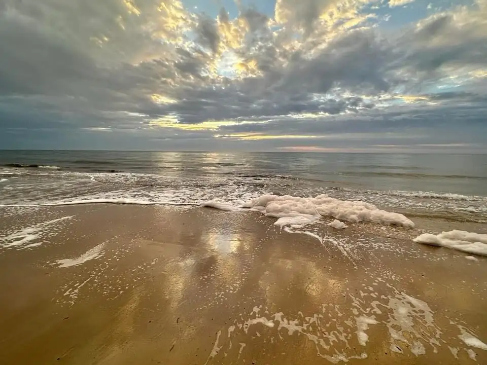 A serene beach scene with gentle waves washing over wet sand under a dramatic, cloudy sky with golden sunlight breaking…