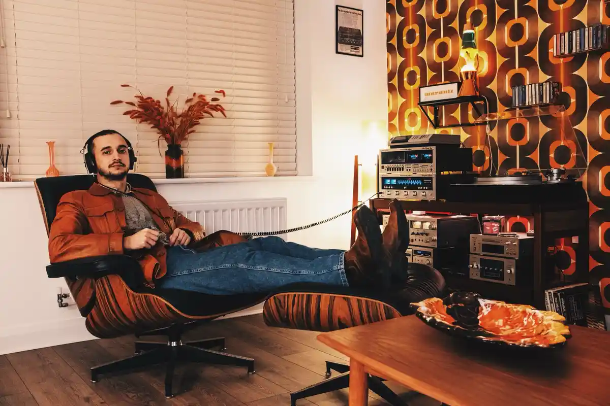 A man lounges in a retro-style wooden lounge chair with his feet up on a matching ottoman, wearing headphones and looking…