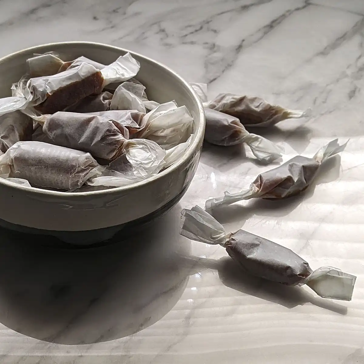 A white ceramic bowl filled with individually wrapped brown candies sits on a white marble surface, with several more…