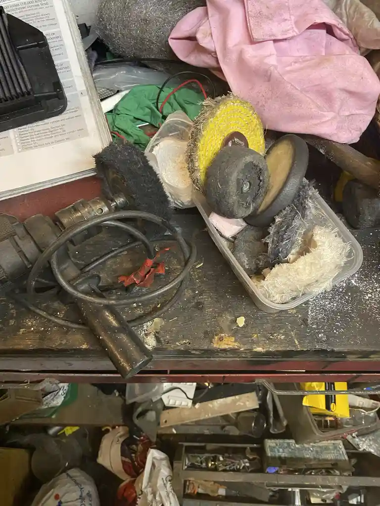 A cluttered workbench is filled with dirty, worn-out grinding wheels, brushes, and other tools in a plastic bin, next to a…
