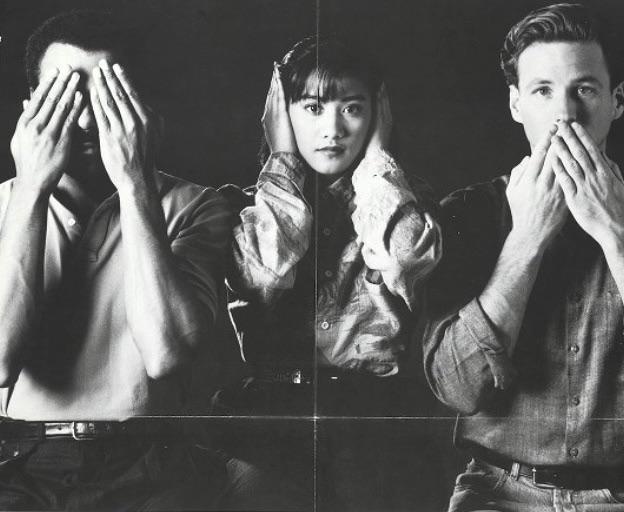 A black-and-white photo shows three people in a row: the person on the left covers their eyes, the person in the middle…