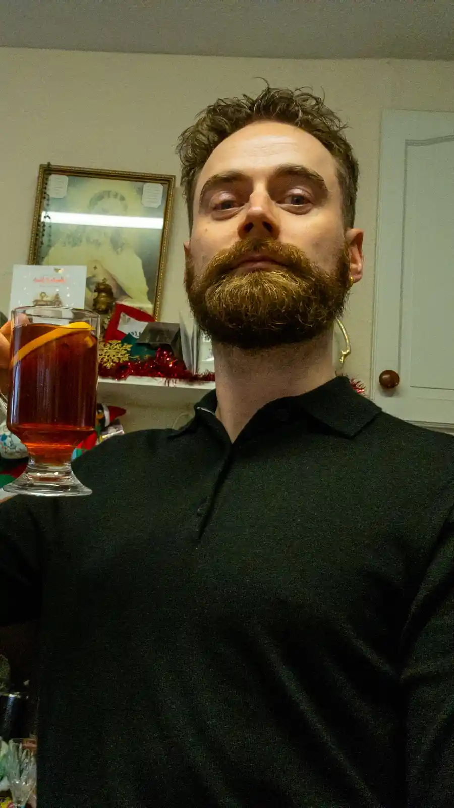 A man with a full beard and dark hair, wearing a black polo shirt, holds up a glass of amber-colored drink with a citrus…