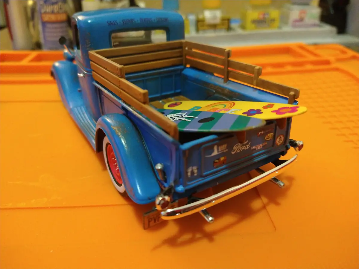 A blue vintage toy pickup truck with wooden bed rails sits on an orange surface, loaded with two longboards decorated in…