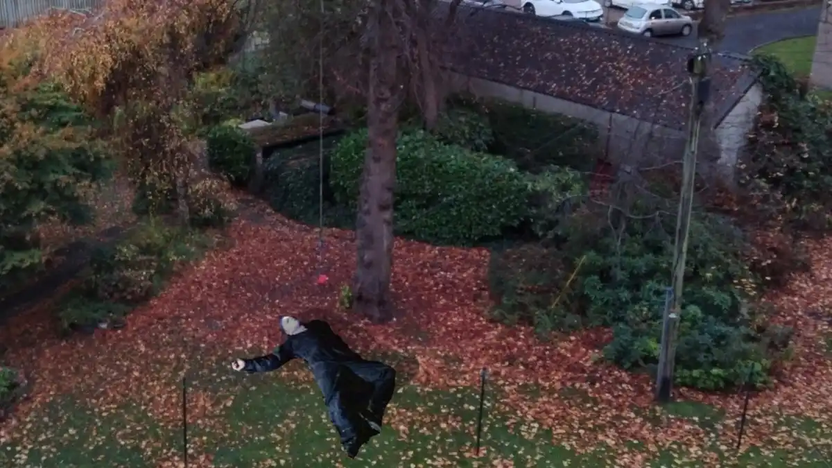 An aerial view of a suburban garden covered in autumn leaves, with a person in a black coat and white face mask digitally…