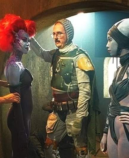 A man in sci-fi armor with glasses and a knit hat stands between two women in futuristic costumes; one has bright red hair…