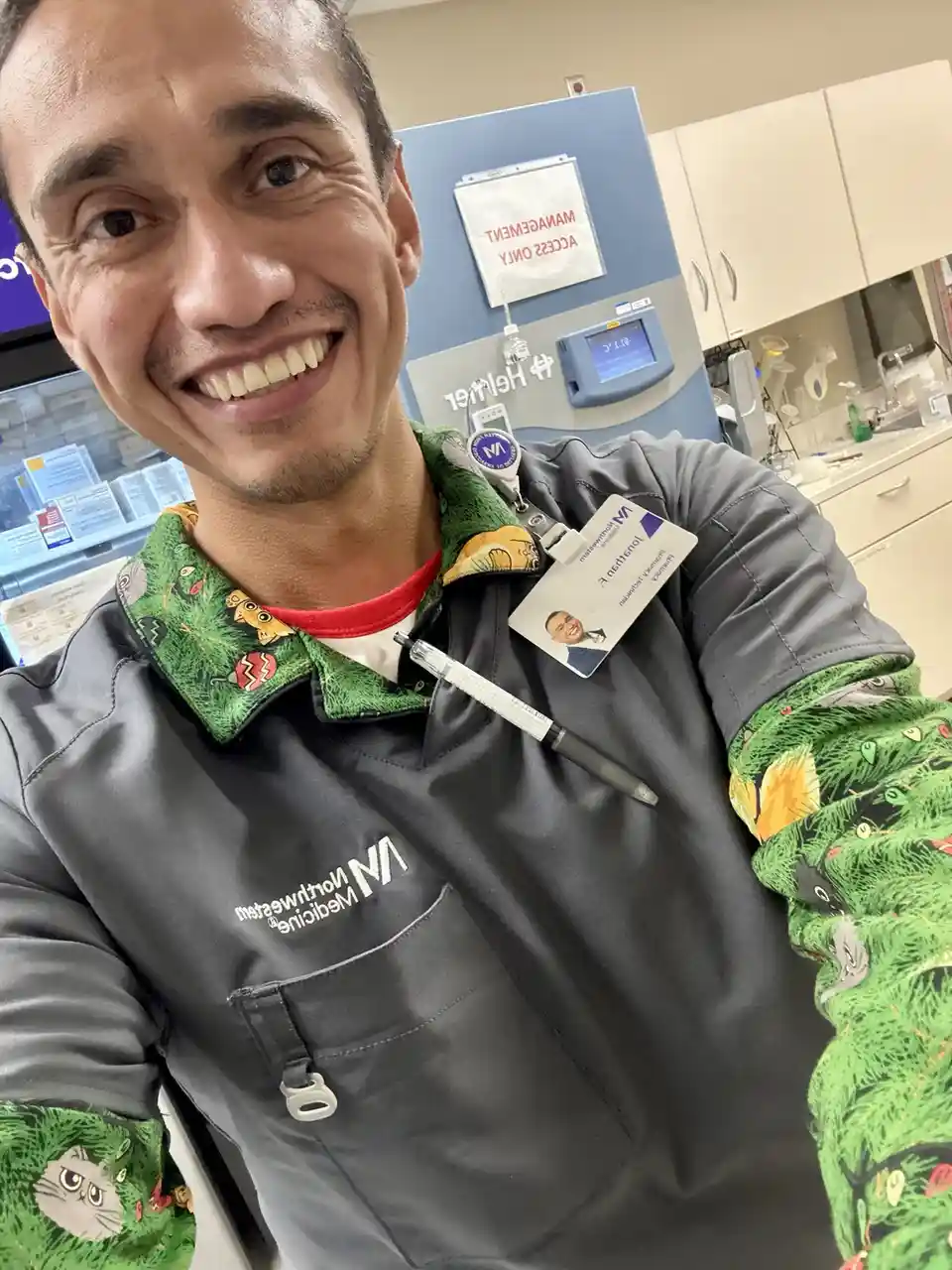 A smiling man in a grey Northwestern Medicine uniform and a colorful green shirt with cartoon cats takes a selfie in a…