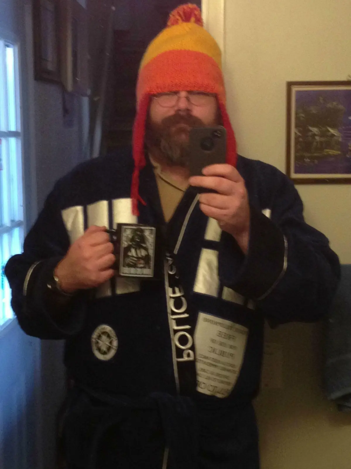 A man with a beard and glasses takes a mirror selfie while wearing a dark blue robe with 'POLICE' written vertically down…