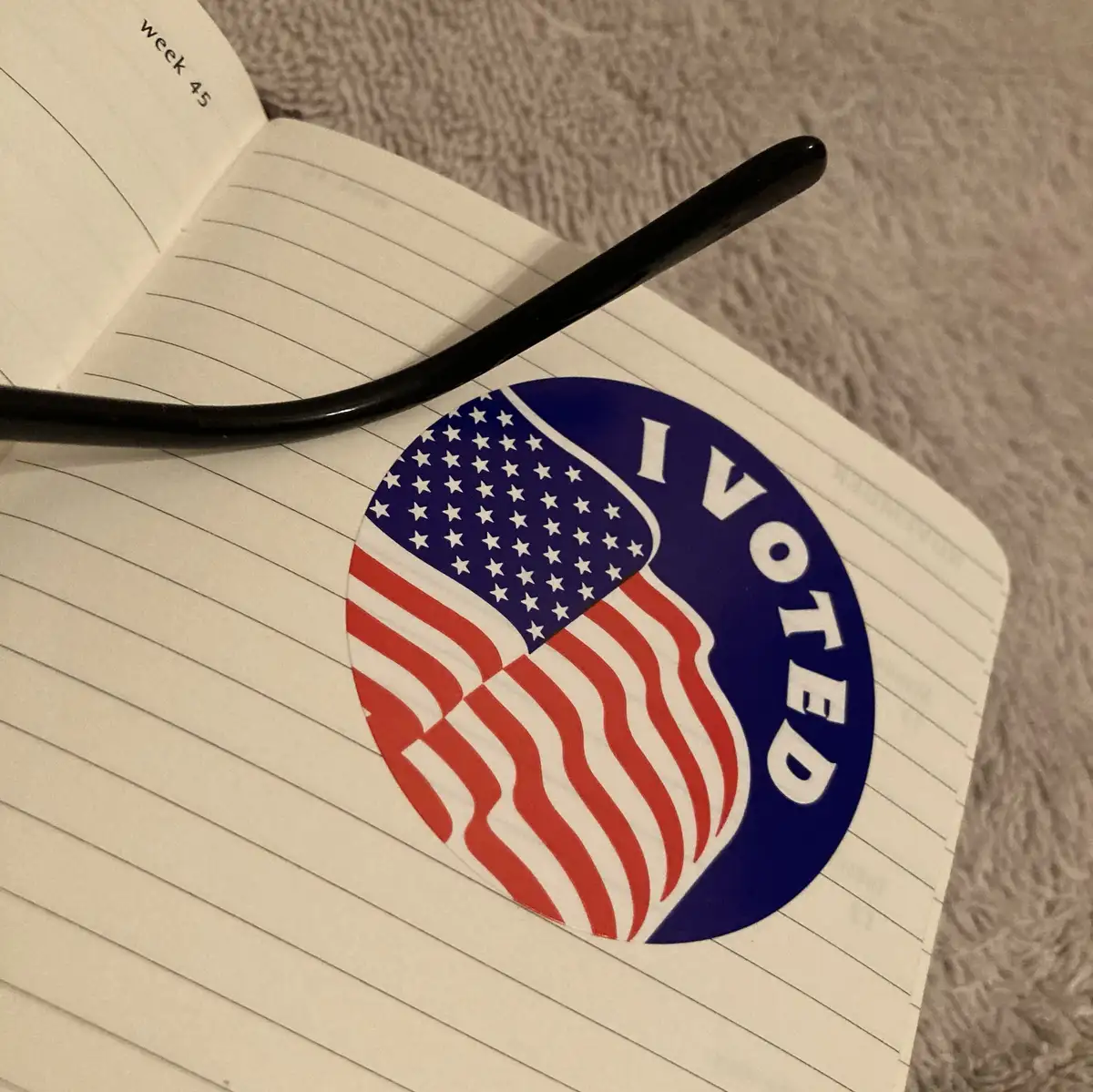 A circular 'I VOTED' sticker featuring the American flag design is placed on a lined notebook page labeled 'week 45'. A…