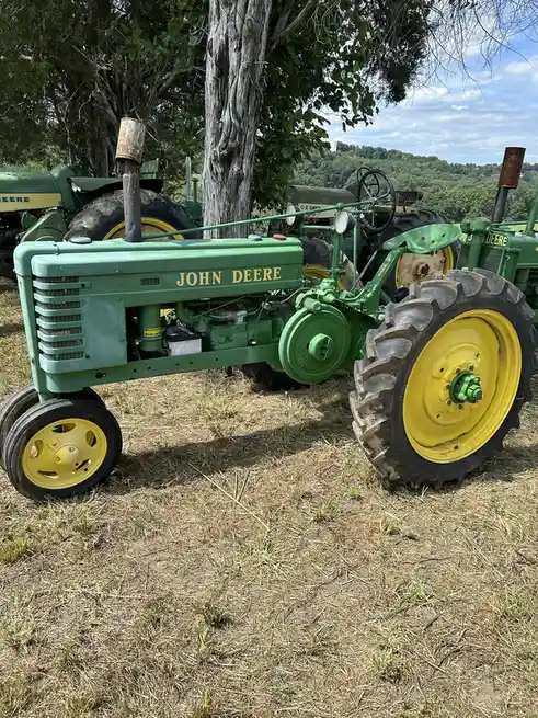 A vintage green and yellow John Deere tractor is parked on dry grass under a tree, with other similar tractors visible in…